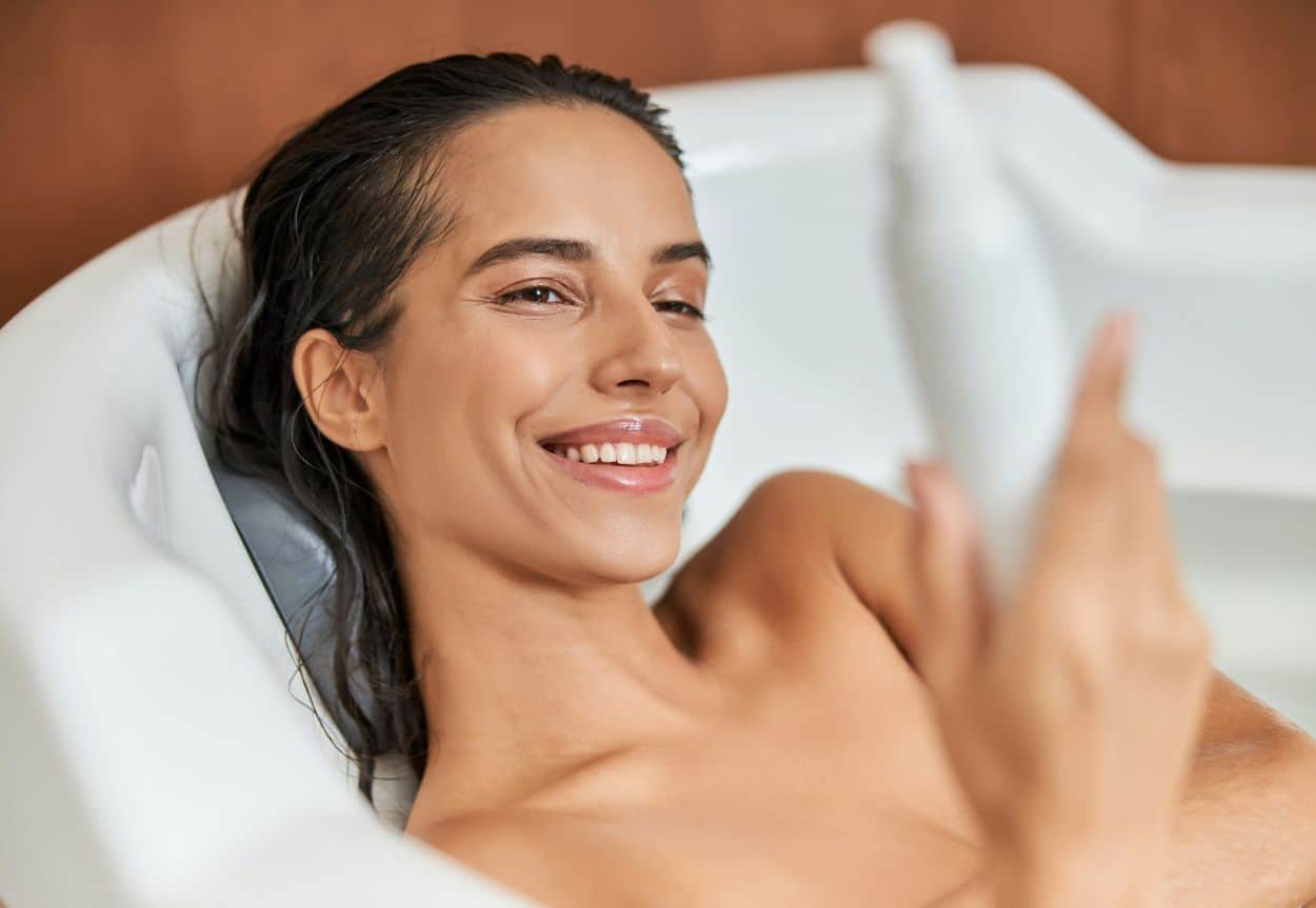 A woman sits in a bathtub holding a white bottle of something and she is smiling at it, like it's colostrum in skincare