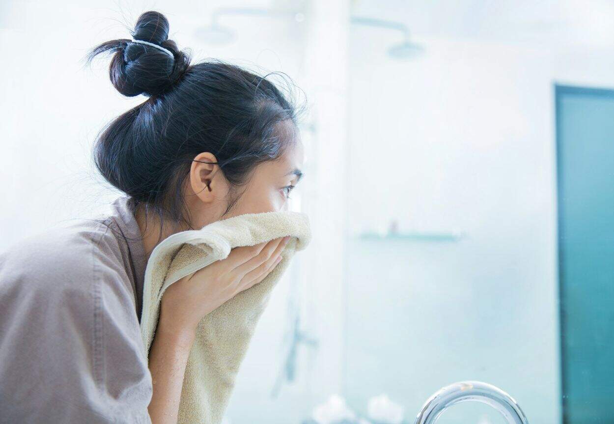A woman pats her face dry at a sink while she looks in a mirror.