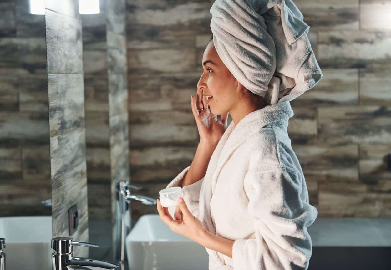 A woman standing at the sink dabbing eye cream under her eyes.